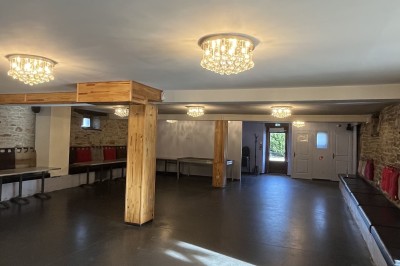 Salle vide avec sol en béton, murs en pierre et bois, tables pliées contre le mur et lustres suspendus au plafond.
