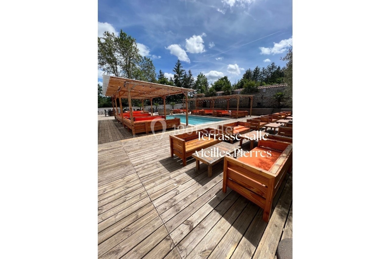 Terrasse en bois avec des tables et banquettes près d'une piscine, entourée de végétation et d'un mur en pierres.