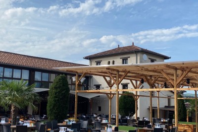 Salle vide avec sol en béton, murs en pierre et bois, tables pliées contre le mur et lustres suspendus au plafond.