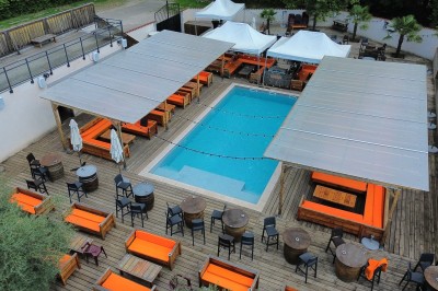 Salle vide avec sol en béton, murs en pierre et bois, tables pliées contre le mur et lustres suspendus au plafond.