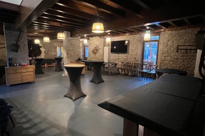 Salle vide avec sol en béton, murs en pierre et bois, tables pliées contre le mur et lustres suspendus au plafond.