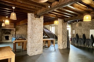 Salle vide avec sol en béton, murs en pierre et bois, tables pliées contre le mur et lustres suspendus au plafond.
