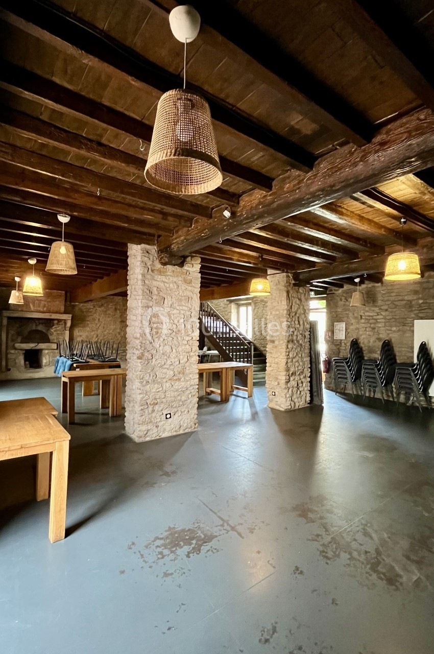 Salle intérieure avec murs en pierre, poutres en bois, tables en bois clair et chaises empilées dans un coin.
