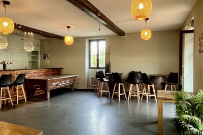 Salle vide avec sol en béton, murs en pierre et bois, tables pliées contre le mur et lustres suspendus au plafond.