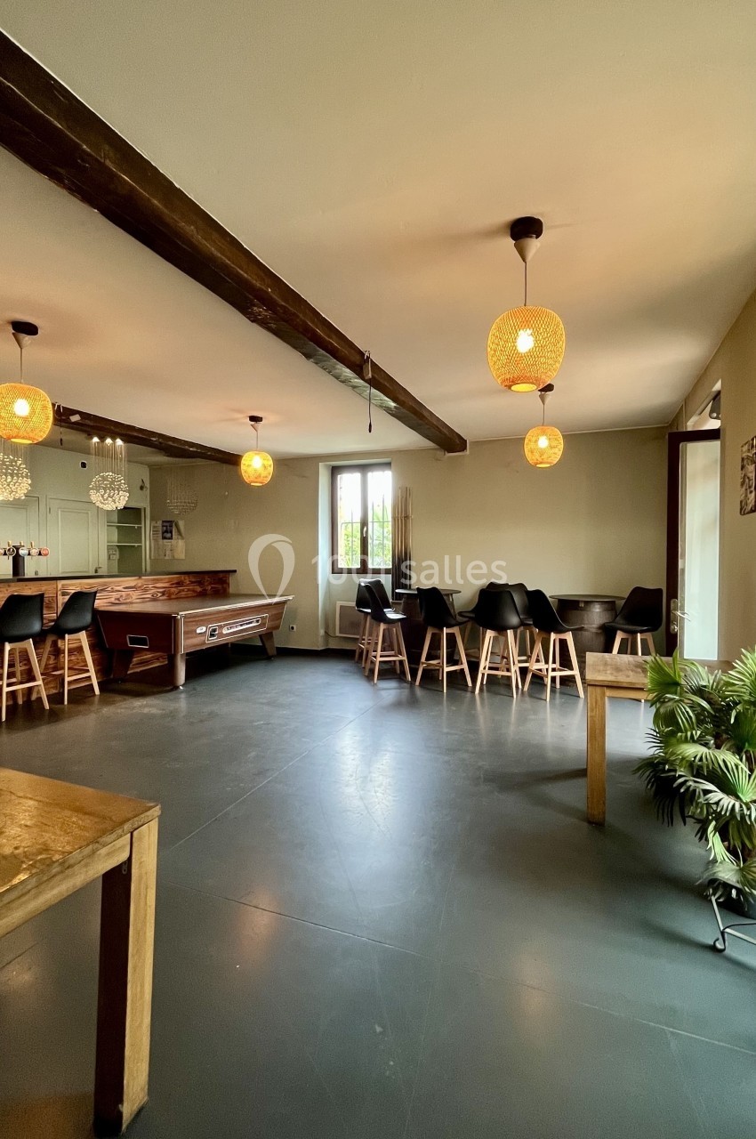 Salle lumineuse avec billard, table en bois, chaises noires, plantes et suspensions en rotin.