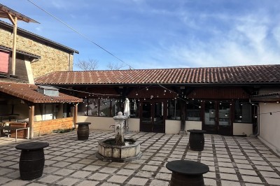 Salle vide avec sol en béton, murs en pierre et bois, tables pliées contre le mur et lustres suspendus au plafond.