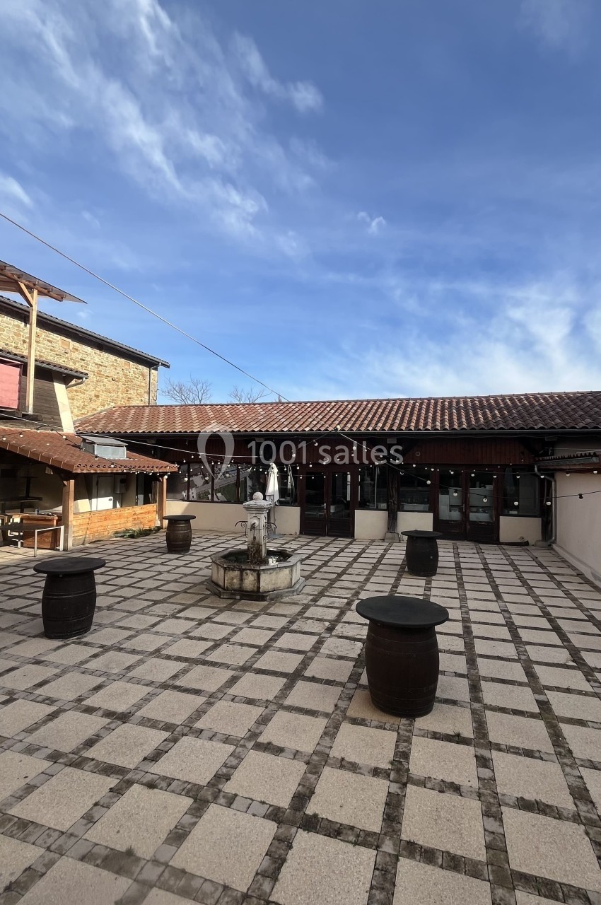 Cour pavée avec une fontaine centrale, entourée de bâtiments en pierre et de tonneaux en bois sous un ciel dégagé.