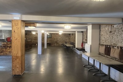 Salle vide avec sol en béton, murs en pierre et bois, tables pliées contre le mur et lustres suspendus au plafond.