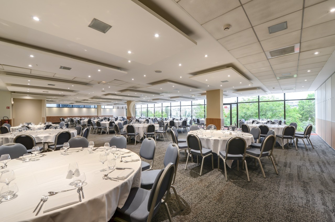 Salle de réception lumineuse avec de grandes baies vitrées, tables rondes dressées et chaises disposées autour.