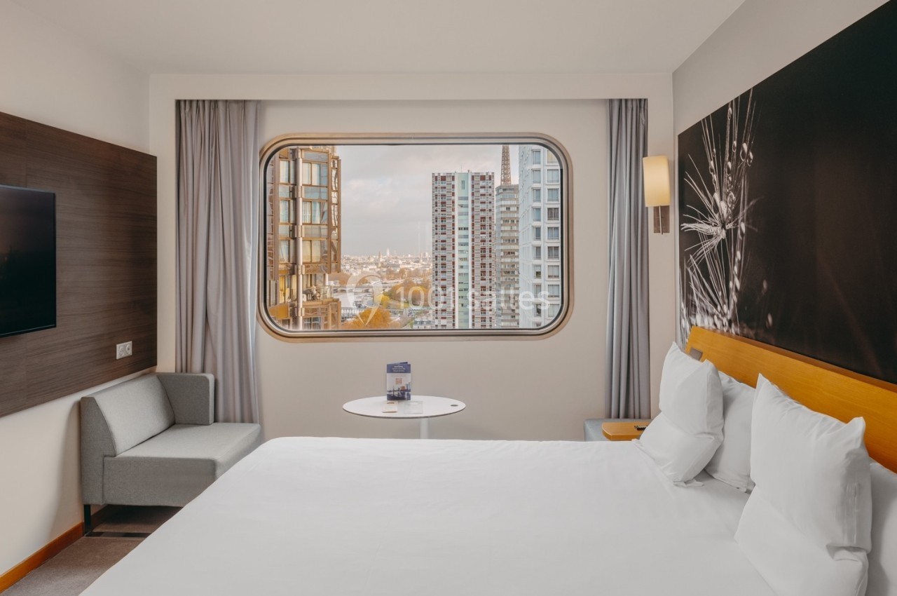 Chambre d'hôtel moderne avec lit double, canapé, table ronde et grande fenêtre offrant une vue sur des immeubles urbains.
