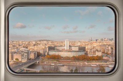 Vue de Paris à travers une fenêtre, montrant la Seine, un bâtiment moderne et l'horizon urbain avec des gratte-ciels.