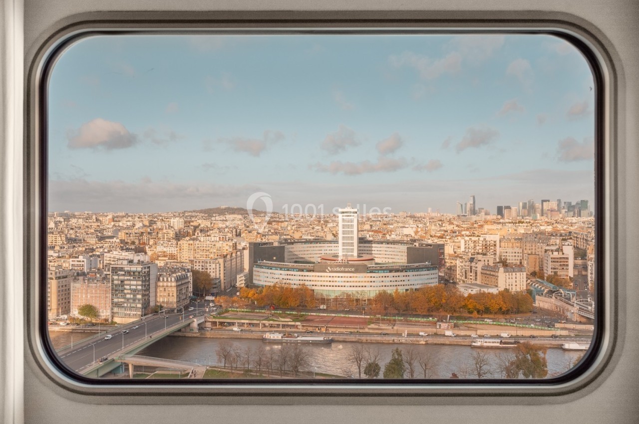 Vue de Paris à travers une fenêtre, montrant la Seine, un bâtiment moderne et l'horizon urbain avec des gratte-ciels.