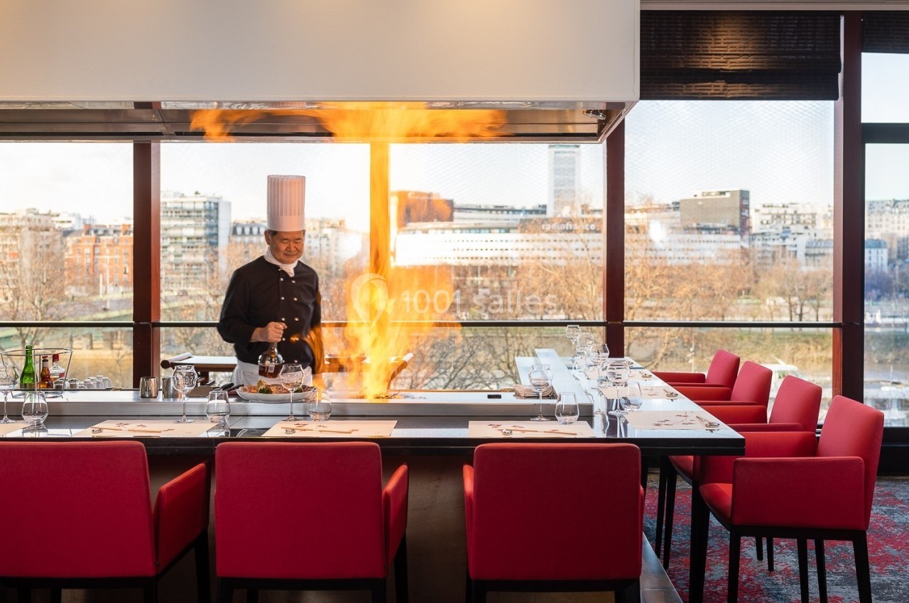 Un chef cuisine devant une flamme vive dans un restaurant avec des tables dressées et des chaises rouges.