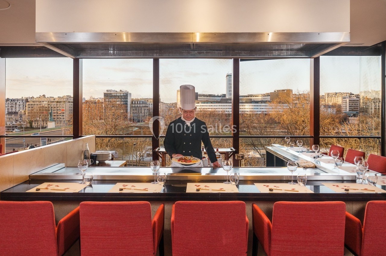 Un chef en tenue blanche présente un plat dans un restaurant avec vue sur une ville à travers de grandes baies vitrées.