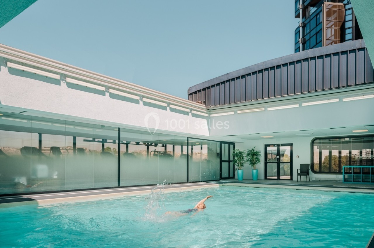 Une personne nage dans une piscine extérieure entourée de bâtiments modernes sous un ciel dégagé.