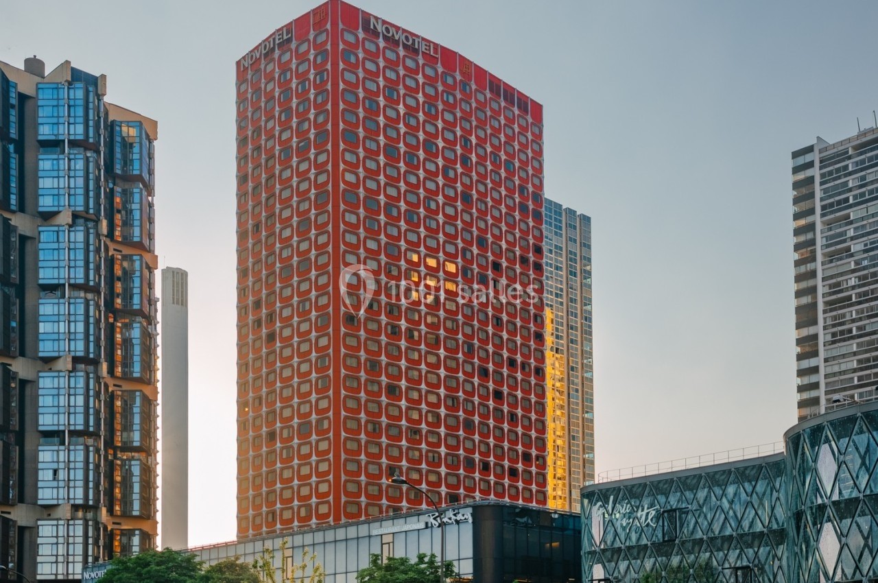 Façade d'un hôtel rouge marqué ’Novotel’, entouré de bâtiments modernes sous un ciel clair.