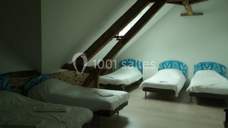 Chambre mansardée avec quatre lits simples, poutres apparentes et lumière naturelle provenant d'une fenêtre.