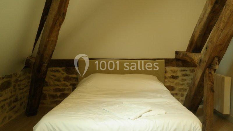 Lit double avec draps blancs dans une chambre mansardée aux poutres apparentes et murs en pierre.