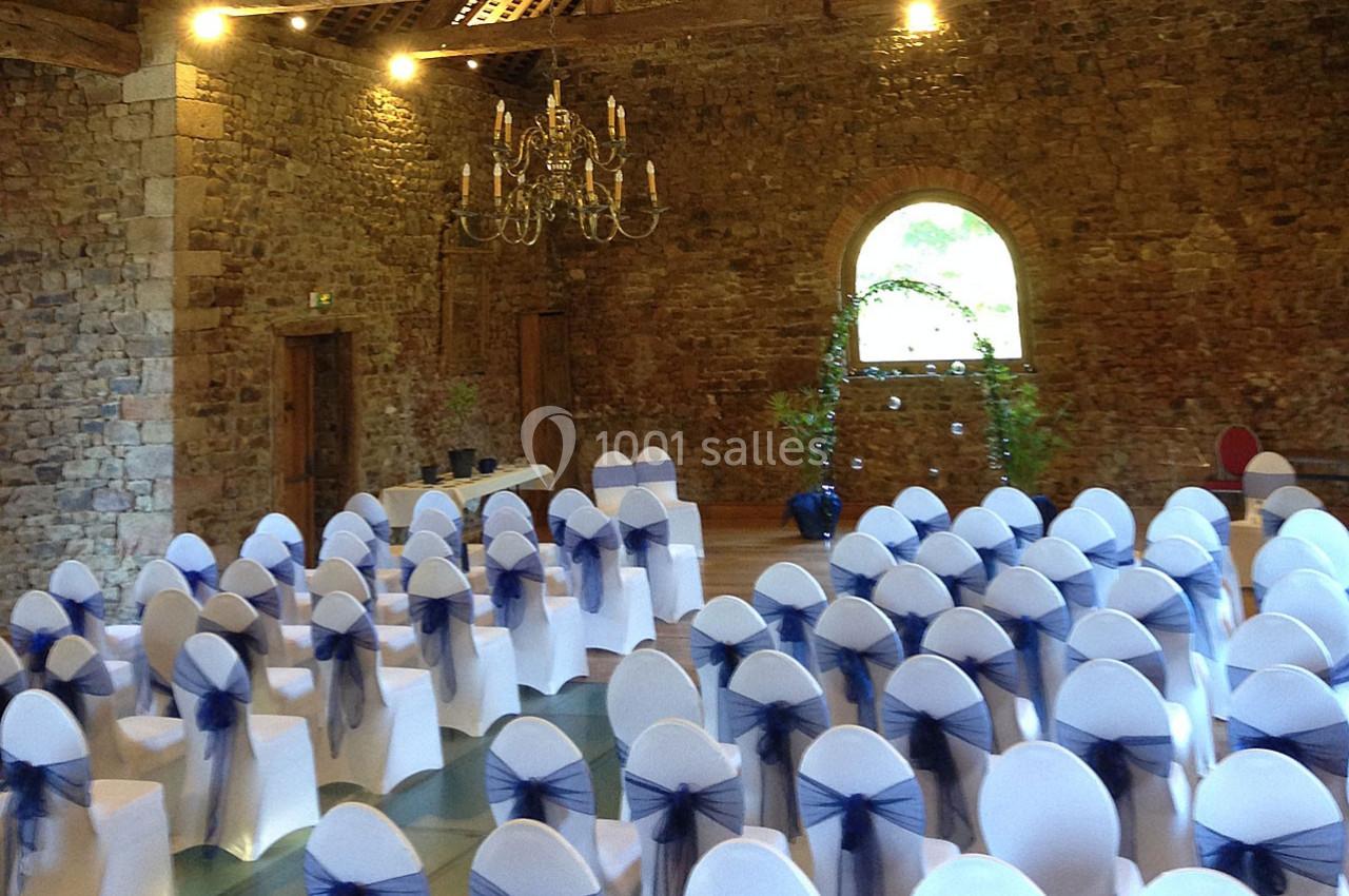 Salle en pierre avec des chaises blanches décorées de nœuds bleus, disposées pour un événement, sous un lustre suspendu.