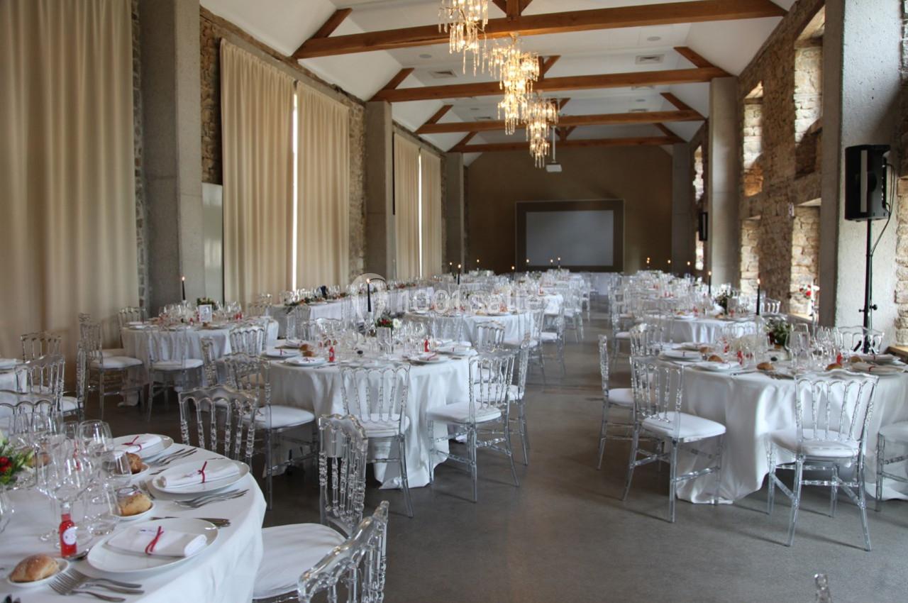 Salle de réception décorée avec des tables rondes dressées, nappes blanches et chaises transparentes, éclairée par des…