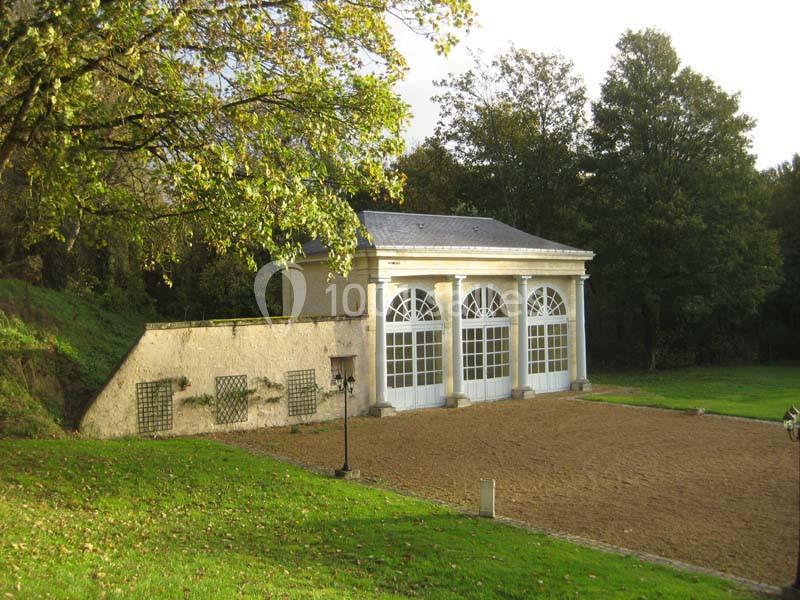 Bâtiment avec grandes fenêtres cintrées, entouré de verdure et d'une cour en gravier, sous un ciel clair.