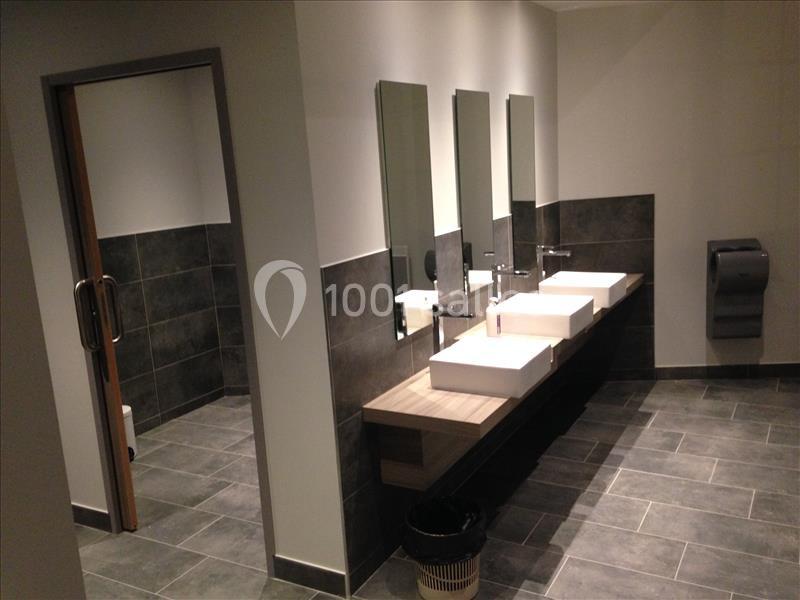 Salle de bain moderne avec trois lavabos, miroirs alignés, carrelage gris et distributeur de papier mural.