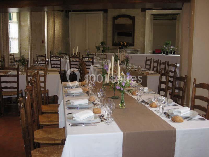 Salle de réception avec tables dressées, nappes blanches, chemins de table beige, vaisselle, verres et décorations florales.