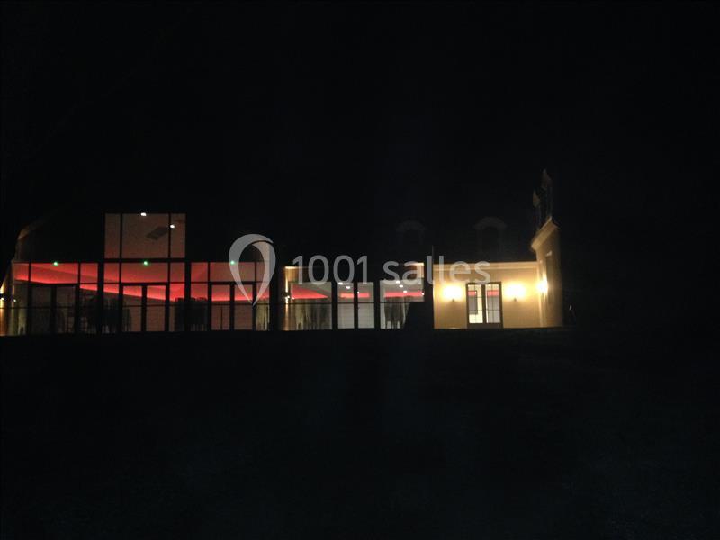 Bâtiment moderne éclairé de nuit avec des lumières rouges et jaunes visibles à travers les grandes fenêtres.