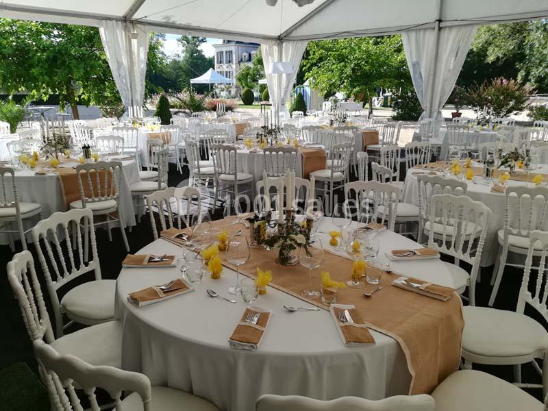 Tables rondes décorées avec nappes blanches, chemins de table beige et serviettes jaunes, sous une tente en extérieur.