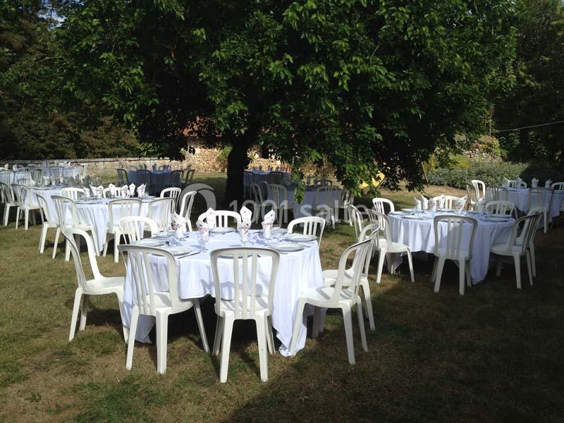 Tables rondes dressées avec nappes blanches et chaises en plastique disposées dans un jardin ombragé.