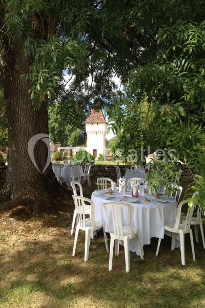 Tables rondes dressées avec nappes blanches sous un grand arbre, dans un jardin avec une tour en arrière-plan.