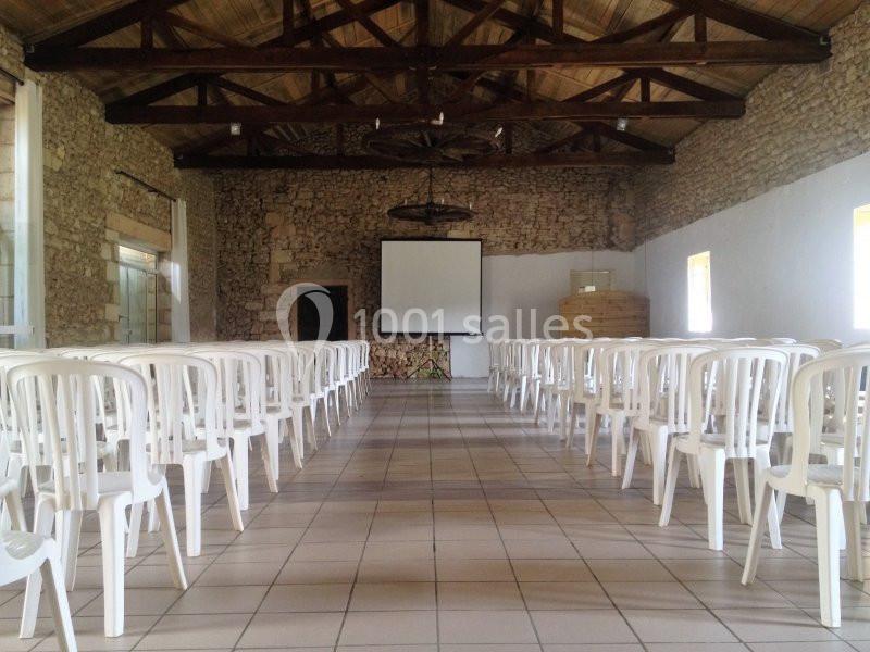 Salle en pierre avec plafond en bois, rangées de chaises blanches en plastique et écran de projection à l'avant.