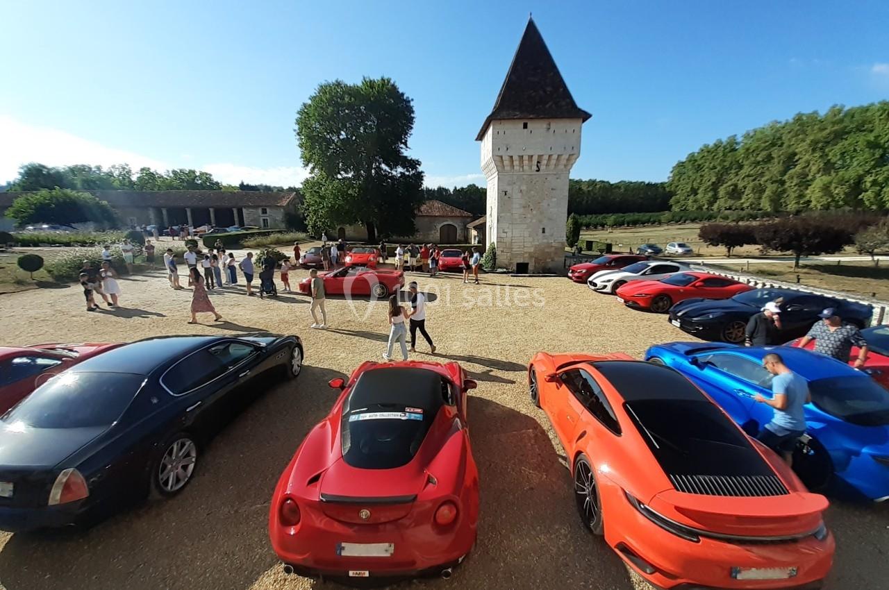 Plusieurs voitures de sport colorées stationnées devant une tour en pierre dans un cadre champêtre par temps ensoleillé.
