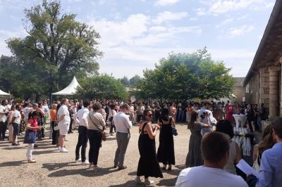 Vue aérienne de plusieurs bus stationnés près d'un bâtiment ancien, entourés de groupes de personnes et de verdure.
