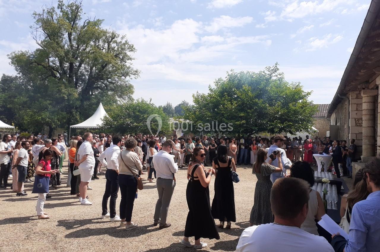 Groupe de personnes rassemblées en extérieur dans une cour ensoleillée, entourée d'arbres et de tentes blanches.