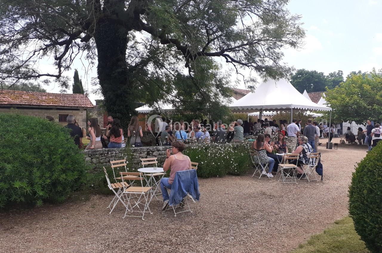 Des personnes assises et debout dans un jardin avec des tables, des chaises et une tente blanche en arrière-plan.
