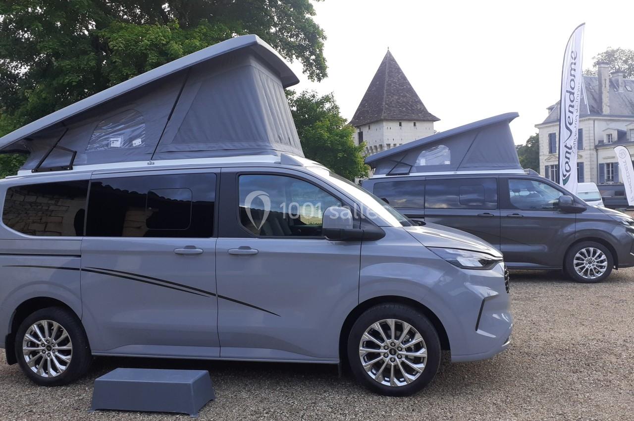 Deux vans gris avec toits relevables stationnés sur un terrain gravillonné devant un bâtiment ancien.