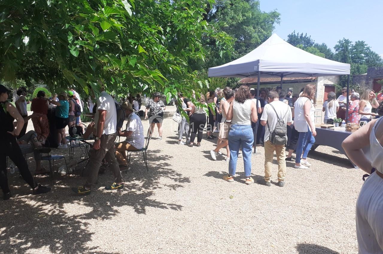Groupe de personnes rassemblées dans un espace extérieur ombragé avec des stands et des tables sous un ciel ensoleillé.