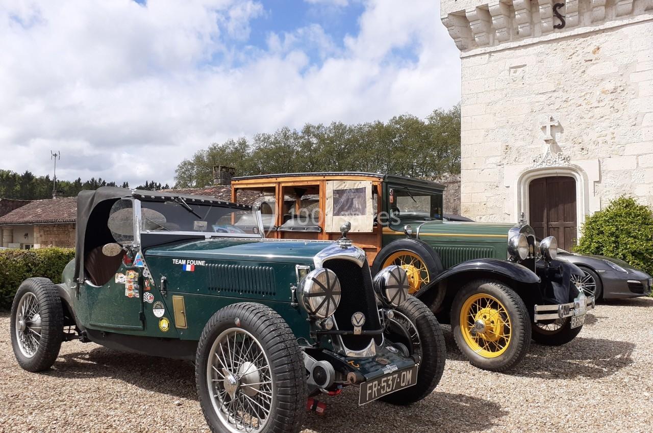 Deux voitures anciennes, une décapotable verte et une berline, stationnées devant un bâtiment en pierre.