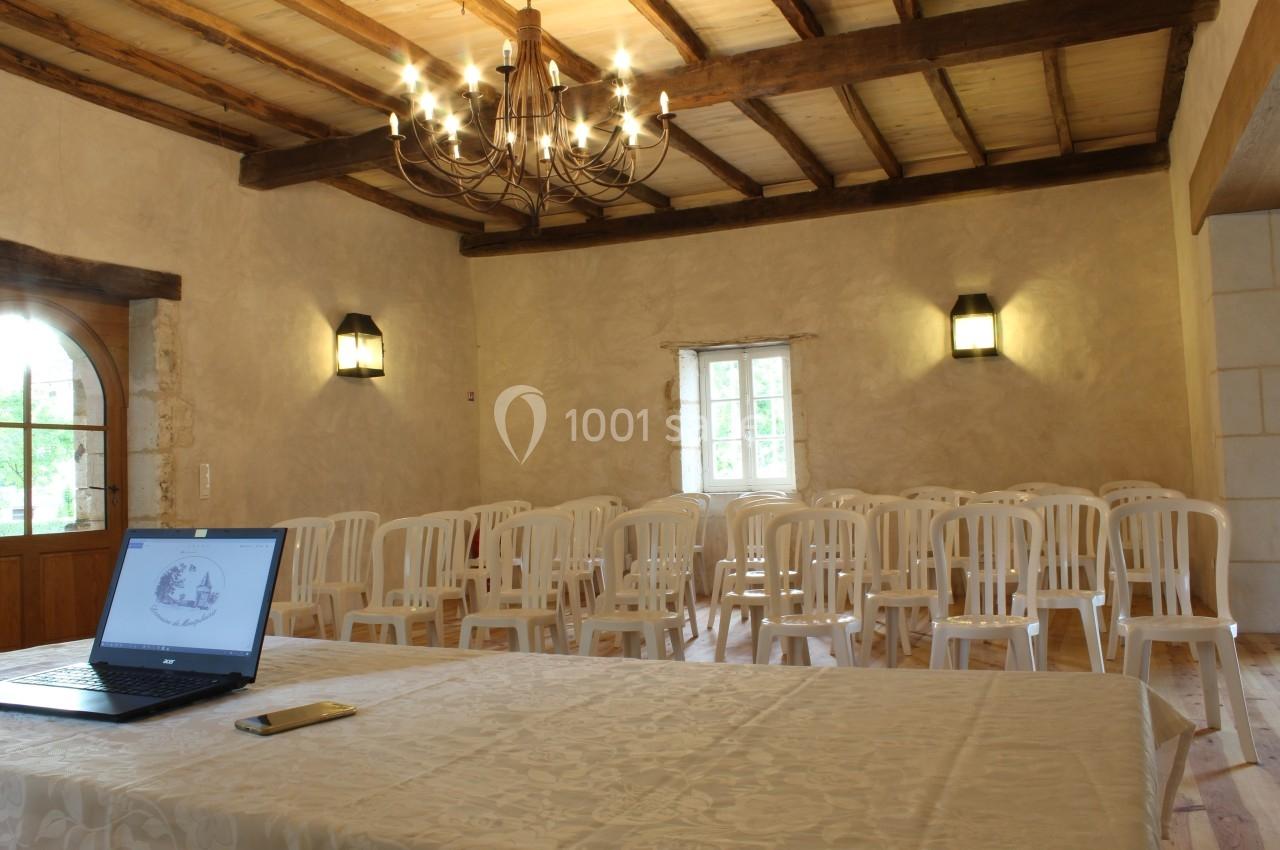Salle lumineuse avec des chaises blanches alignées, une table recouverte d'une nappe et un ordinateur portable ouvert.