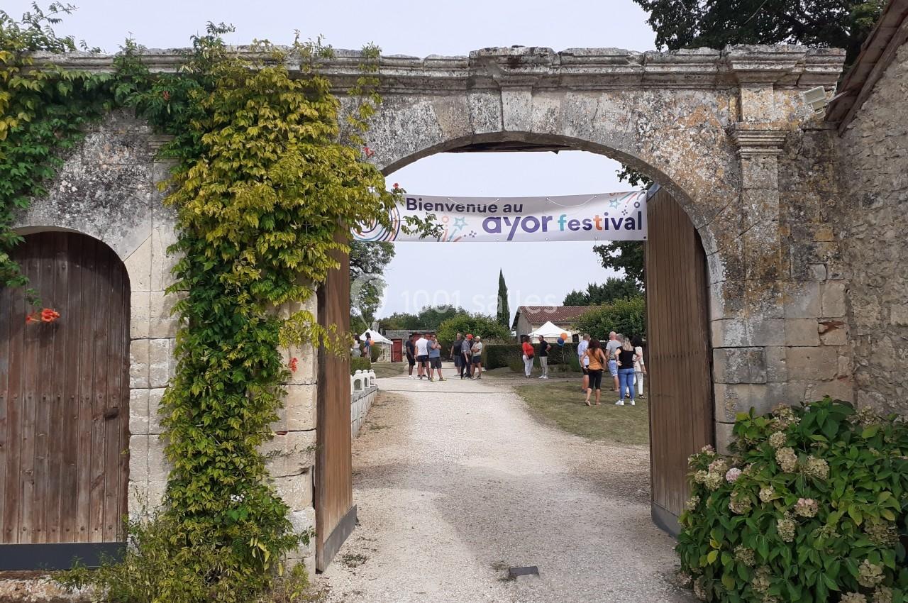 Entrée en pierre avec une banderole ’Bienvenue au AYOR festival’, menant à un chemin où des personnes se rassemblent.