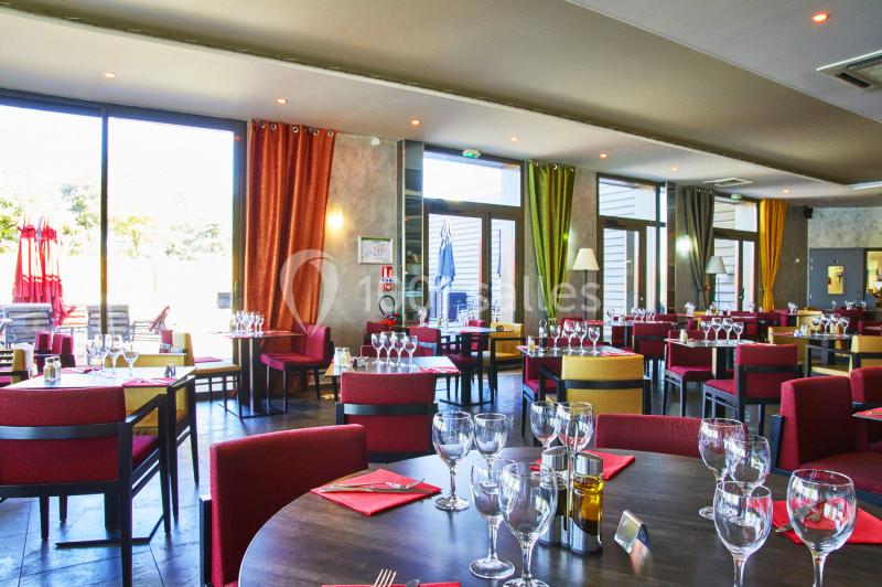 Salle de restaurant lumineuse avec tables dressées, nappes rouges, grandes baies vitrées et vue sur une terrasse.
