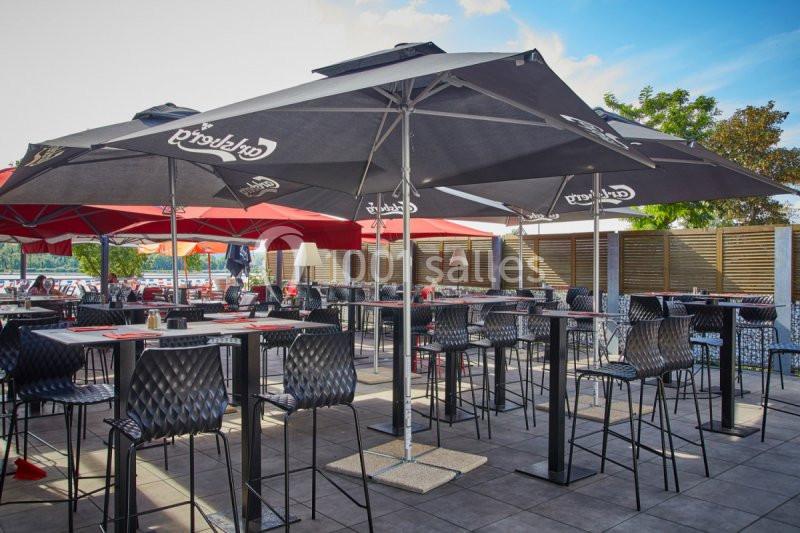 Terrasse extérieure avec tables, chaises hautes et parasols noirs, entourée de clôtures et végétation en arrière-plan.