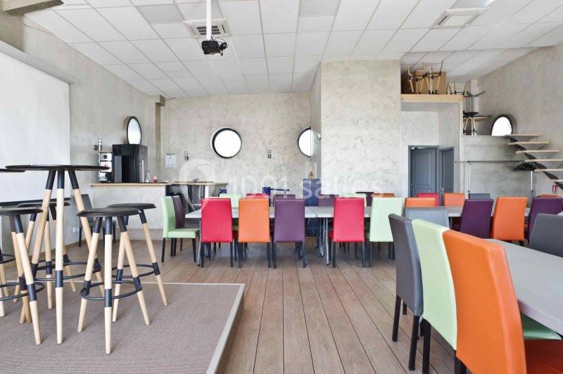 Salle lumineuse avec des tables et des chaises colorées, un bar en arrière-plan et un plafond avec des spots encastrés.