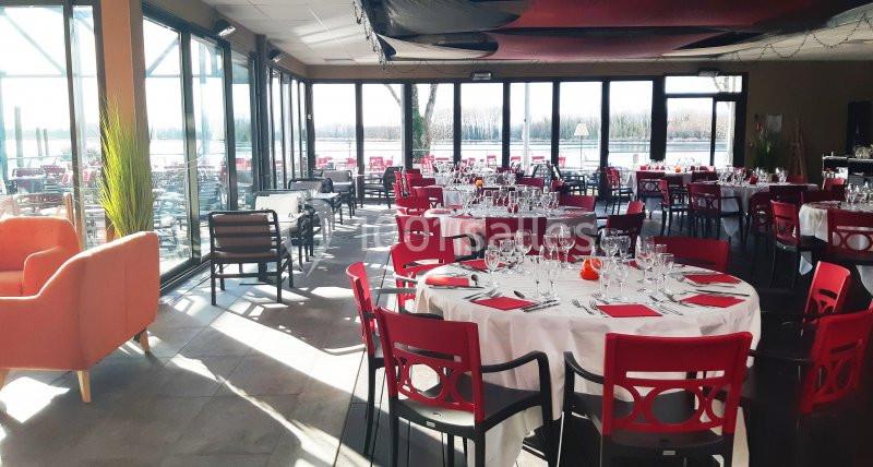 Salle de restaurant lumineuse avec grandes baies vitrées, tables dressées et chaises rouges.