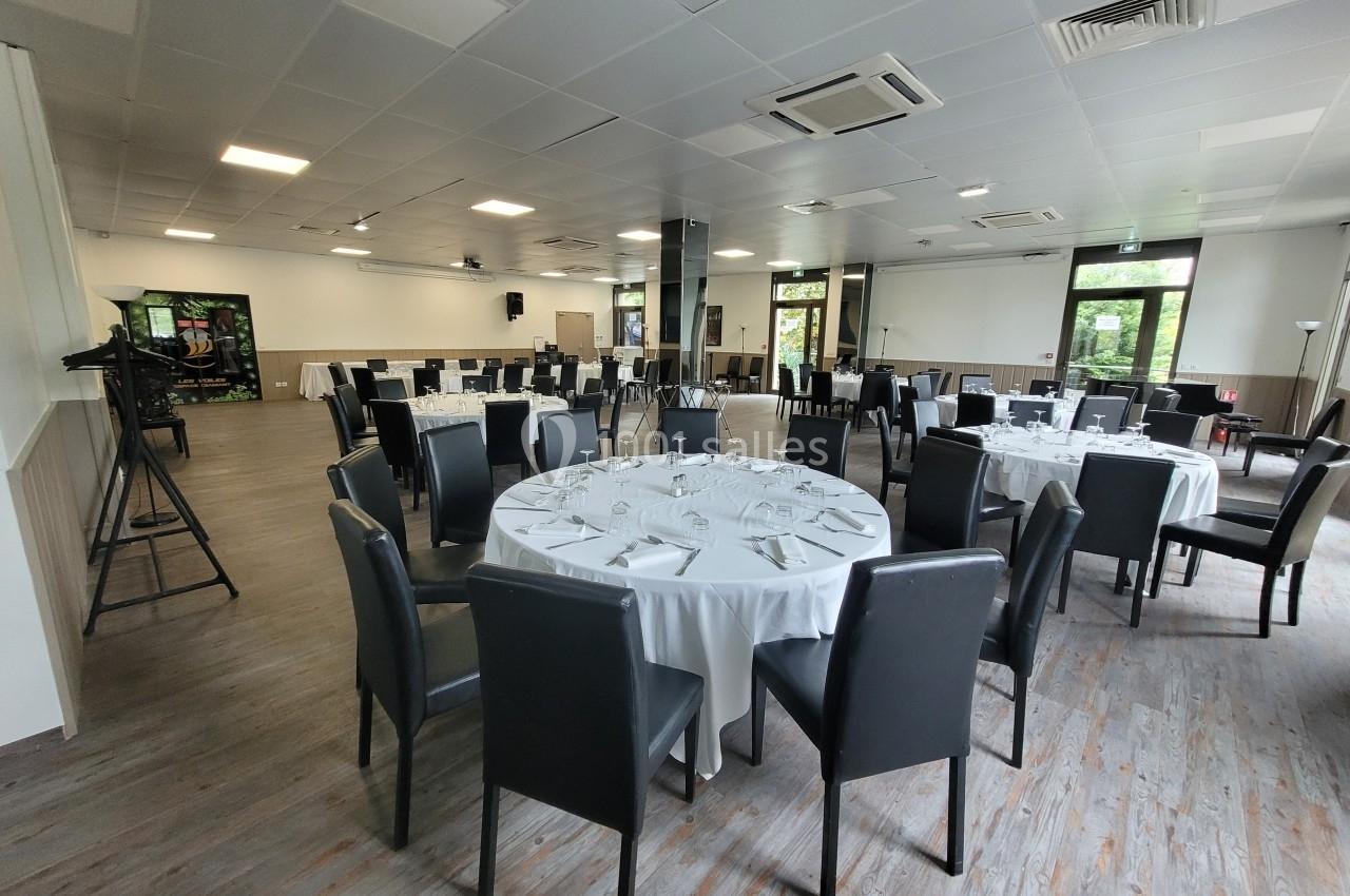 Salle de réception lumineuse avec tables rondes dressées, chaises noires et grandes baies vitrées donnant sur l'extérieur.