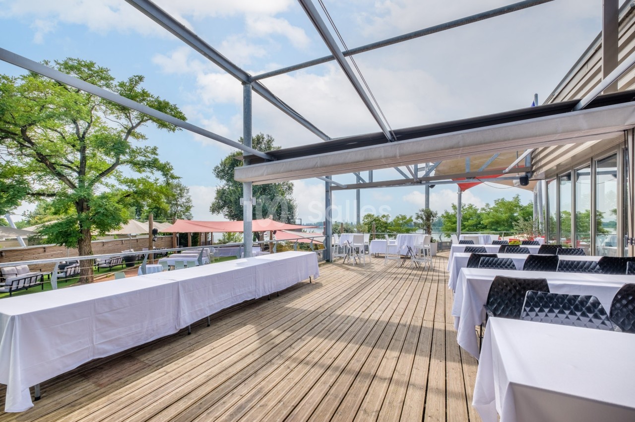 Terrasse en bois aménagée avec des tables dressées, entourée de verdure et partiellement abritée par une structure…