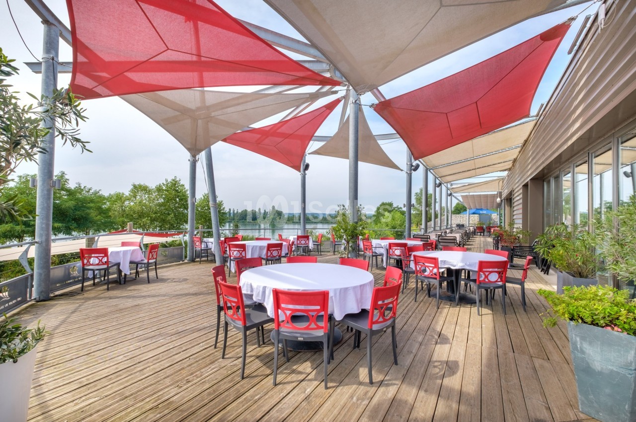 Terrasse en bois avec tables rondes dressées, chaises rouges et grandes toiles d'ombrage, vue sur un paysage verdoyant.