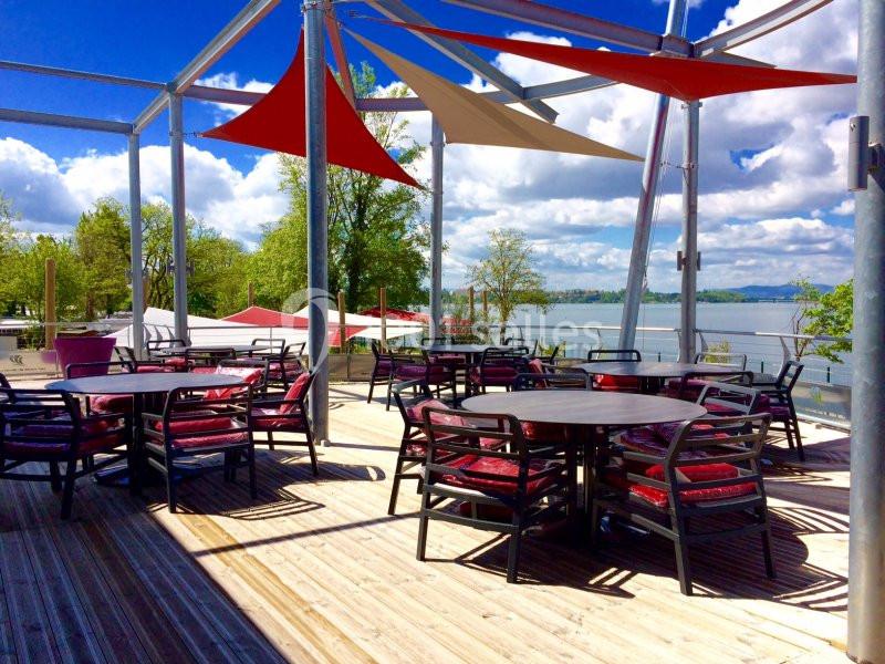 Terrasse en bois avec tables et chaises, surplombant un lac sous un ciel bleu avec des voiles d'ombrage colorées.