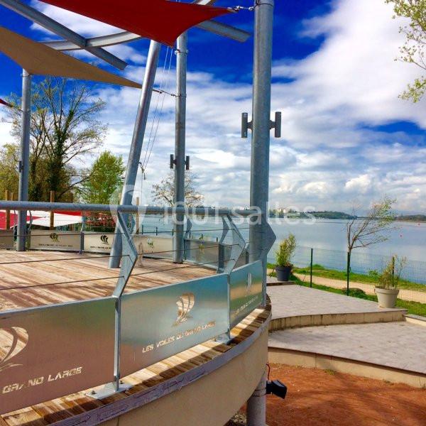 Terrasse en bois avec garde-corps métalliques, surplombant un lac entouré d'arbres sous un ciel partiellement nuageux.